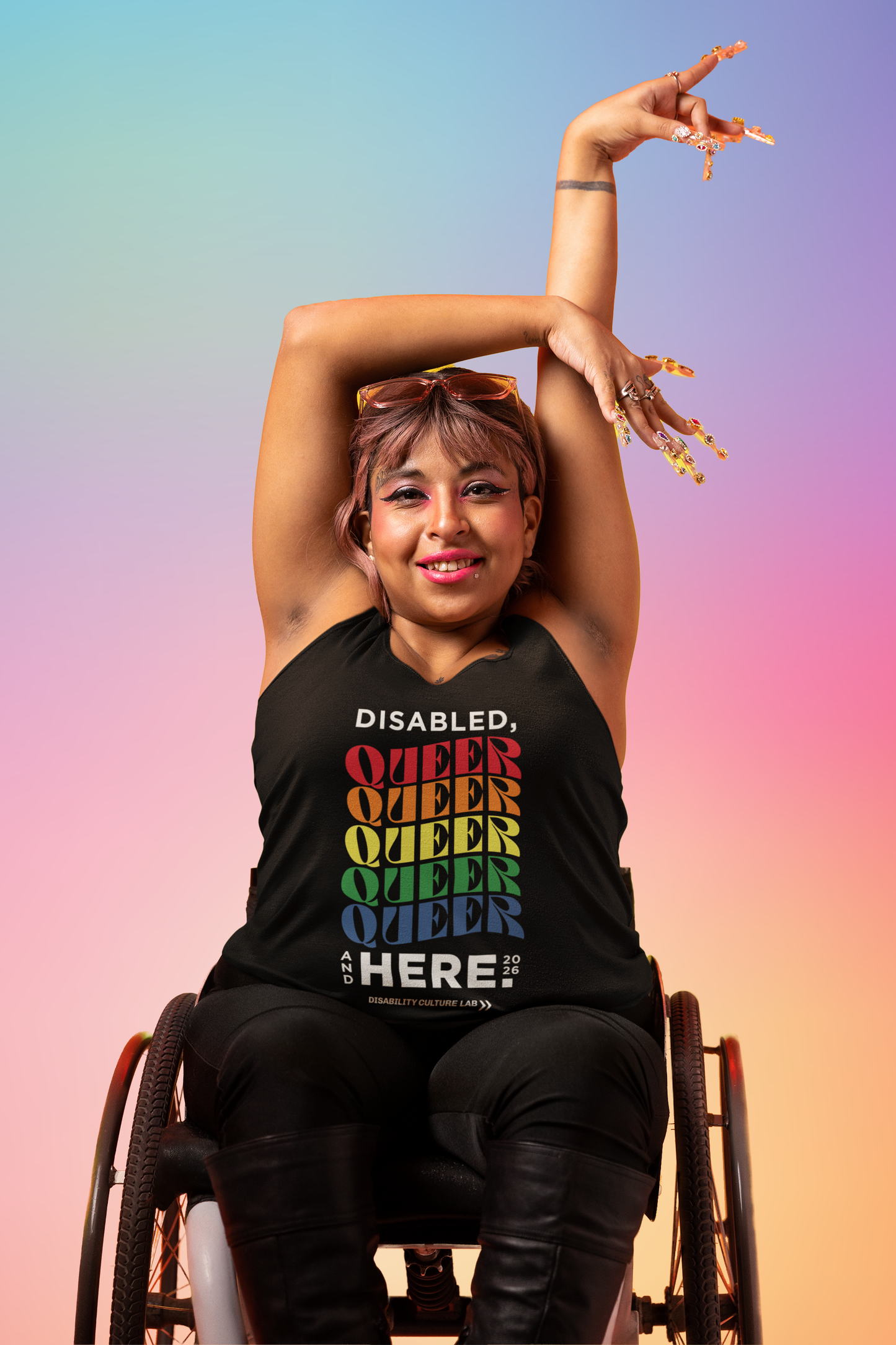 A person with light brown skin sits in a wheelchair, their hands above their head. They're wearing black pants, boots, and a muscle tank that reads, "Disabled, Queer, and Here 2026." "Disabled" is in black, and then "Queer" is written five times in a rainbow of colors, with a curve to the letters. Below that is black text that says "And Here." The "And" is written in small text vertically next to the "Here" which is the same size as "Disabled" and "Queer." To the right of that is "2026" in small text.
