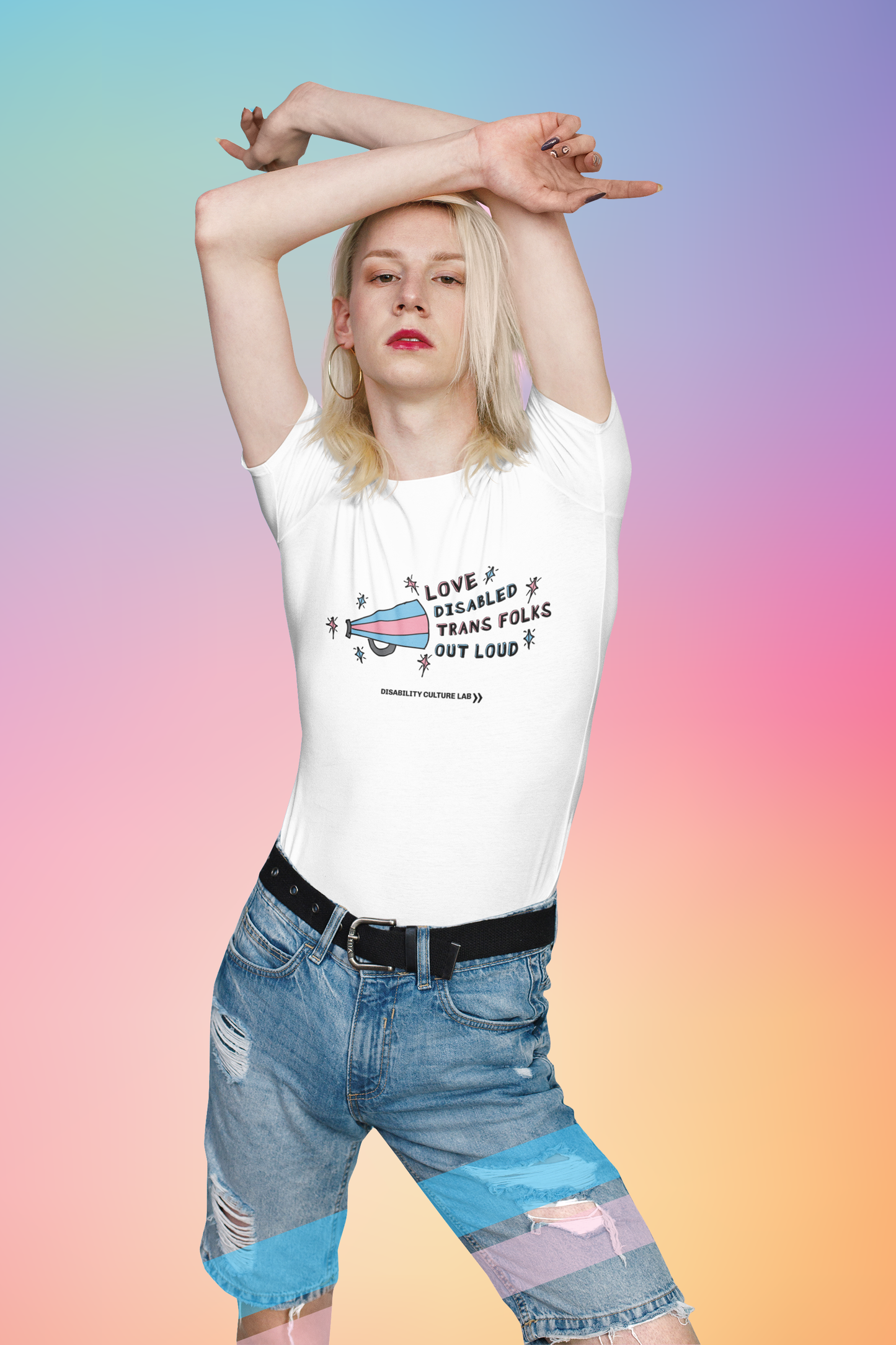 A white person with shoulder length blonde hair. Their arms are up behind their head and they are wearing blue jean shorts and a white short sleeve T-shirt with a striped light blue and light pink megaphone on the left side and alternating pink and blue text outlined in black that reads, "Love Disabled Trans Folks Out Loud." There are light pink and light blue sparkles outlined in black surrounding the megaphone and the text. Below the design is the black Disability Culture Lab logo.