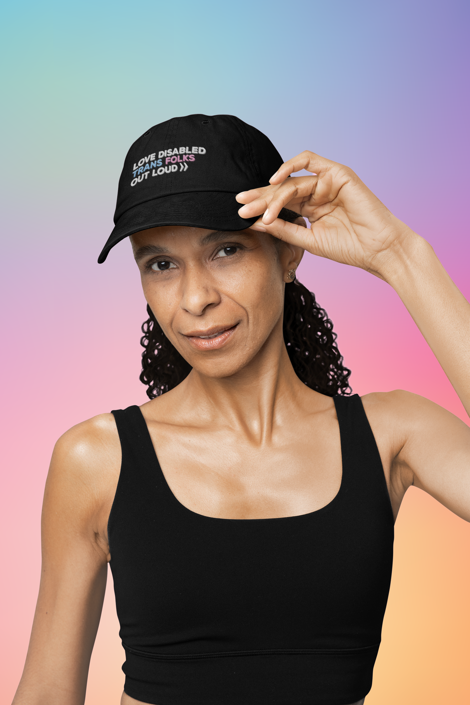 A person with brown skin and long curly black hair that is pulled back. They are wearing a black tank top and a black embroidered hat that reads, "Love Disabled Trans Folks Out Loud." The text is in the colors of the trans flag, with "Love Disabled" in white, "Trans" in light blue, "Folks" in light pink, and "Out Loud" in white. Next to the words "Out Loud" are two white arrows, from the Disability Culture Lab logo.