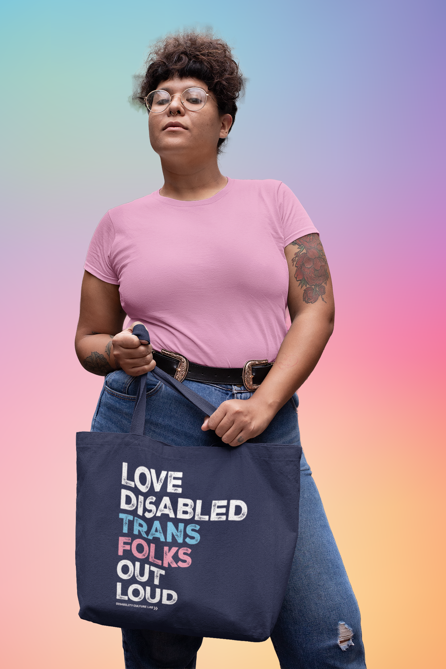 A person with light brown skin, glasses, and brown hair that is pulled up. They are wearing a pink T-shirt and blue jeans. They are holding a navy blue tote bag that reads, "Love Disabled Trans Folks Out Loud." The text is in the colors of the trans flag, with "Love Disabled" in white, "Trans" in light blue, "Folks" in light pink, and "Out Loud" in white. Below the design is the white Disability Culture Lab logo.