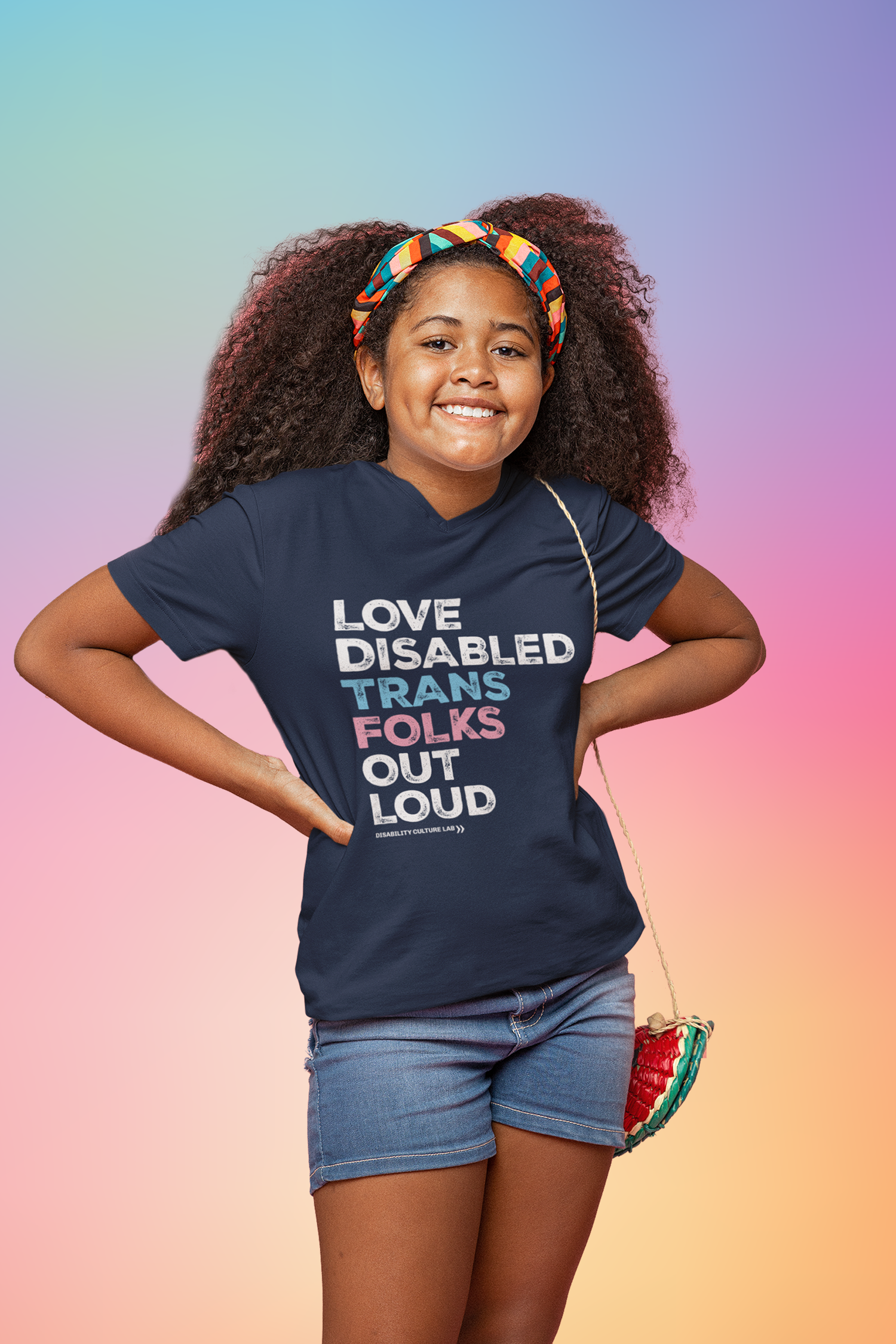 A young Black person with long curly brown hair and a headband. They have a watermelon bag and are wearing blue shorts and a navy blue short sleeve T-shirt that reads, "Love Disabled Trans Folks Out Loud." The text is in the colors of the trans flag, with "Love Disabled" in white, "Trans" in light blue, "Folks" in light pink, and "Out Loud" in white. Below the design is the white Disability Culture Lab logo.