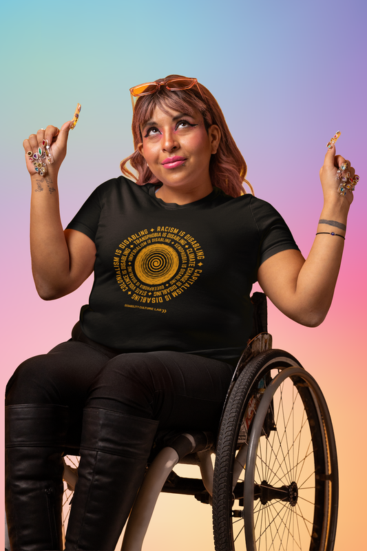 A person with light brown skin and shoulder length hair sits in a wheelchair wearing the black "Oppression Is Disabling" t-shirt, with black pants, long black boots, and yellow sunglasses. Their shirt has a circular design in the middle. In the center of the design there is an orange spiral, with orange text surrounding it in a circle that reads "Racism is disabling. Capitalism is disabling. Colonialism is disabling. Transphobia is disabling. Climate change is disabling," and more. Below is the DCL logo.