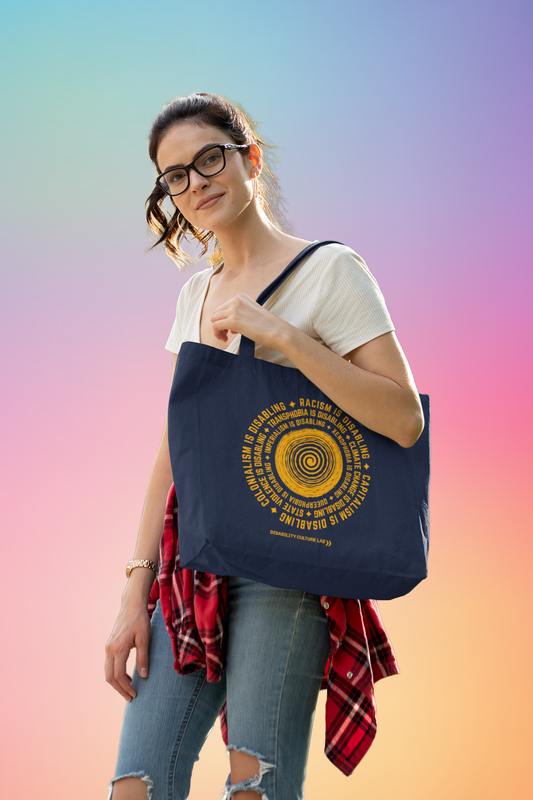 A white person with brown ponytail and glasses, wearing a white T-shirt, ripped jeans, and a red flannel. They are holding a navy blue tote bag with a circular design in the middle. In the center of the design there is an orange spiral, with an orange text surrounding it in a circle that reads, "Racism is disabling. Capitalism is disabling. Colonialism is disabling. Transphobia is disabling. Climate change is disabling. State violence is disabling. Imperialism is disabling. Xenophobia is disabling."