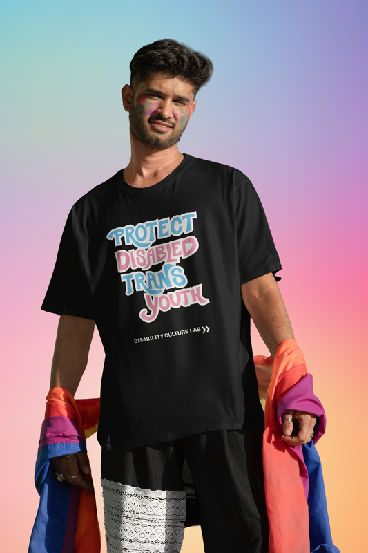 A brown-skinned person with short black hair with rainbow paint on their cheeks, holding a pride flag. They are wearing black pants and a black short sleeve T-shirt that reads “Protect Disabled Trans Youth” in hand drawn letters in the trans pride flag colors (light pink, sky blue, and white). Below is the white Disability Culture Lab logo.