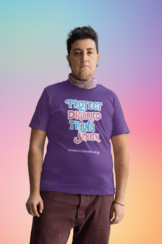A light-skinned person with short brown hair is wearing brown pants and a purple short sleeve T-shirt that reads, “Protect Disabled Trans Youth” in hand drawn letters in the trans pride flag colors (light pink, sky blue, and white). Below is the white Disability Culture Lab logo. They are also wearing a purple bandana around their neck.