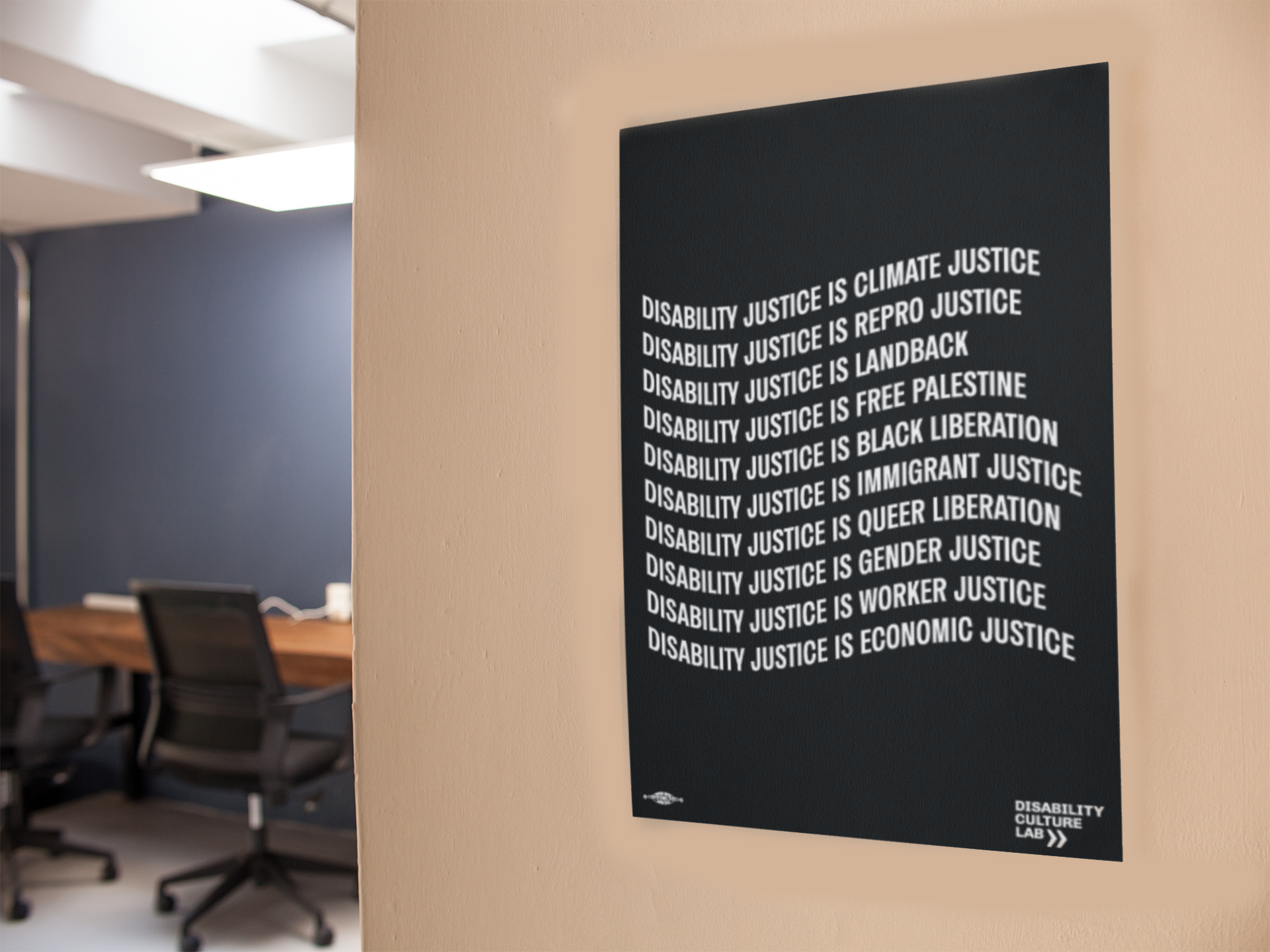 A black poster with the words: "Disability Justice Is... Climate Justice, Repro Justice, Landback, Free Palestine, Black Liberation, Immigrant Justice, Queer Liberation, Gender Justice, Worker Justice, Economic Justice" in a white frame on a beige wall. Black office chairs, a blue wall, and brown desk are in the background.