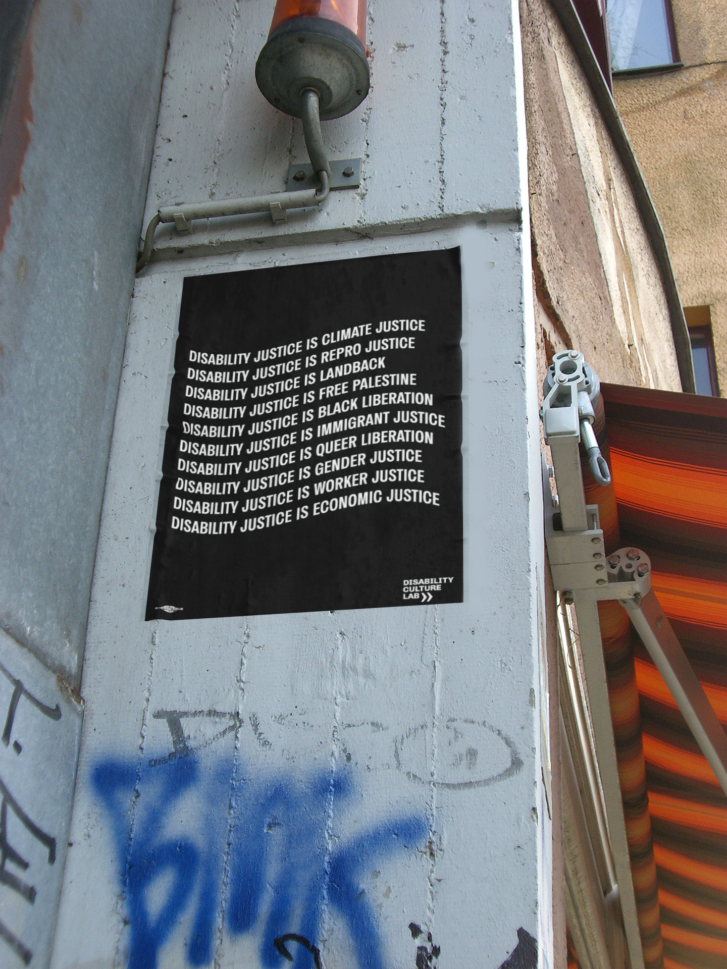 A black poster with the words: "Disability Justice Is... Climate Justice, Repro Justice, Landback, Free Palestine, Black Liberation, Immigrant Justice, Queer Liberation, Gender Justice, Worker Justice, Economic Justice" on a post outside that also features blue and black grafitti and an orange awning. White Disability Culture Lab logo.