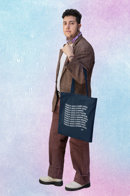 A person with short curly hair, brown slacks, and a brown blazer wearing a suit jacket and a navy tote bag with the words: "Disability Justice Is... Climate Justice, Repro Justice, Landback, Free Palestine, Black Liberation, Immigrant Justice, Queer Liberation, Gender Justice, Worker Justice, Economic Justice."