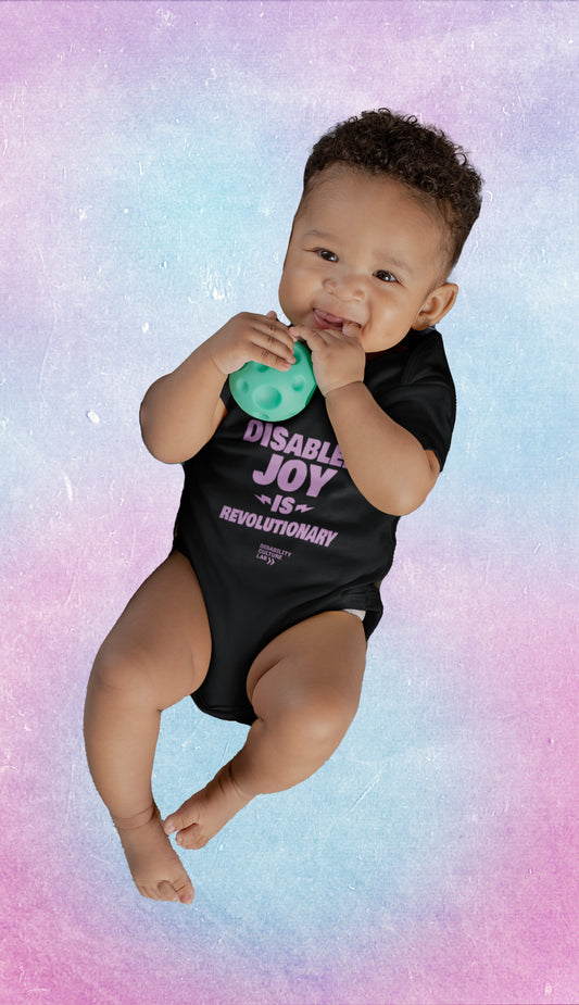 A brown-skinned baby wears a black onsie that says "Disabled Joy is Revolutionary."