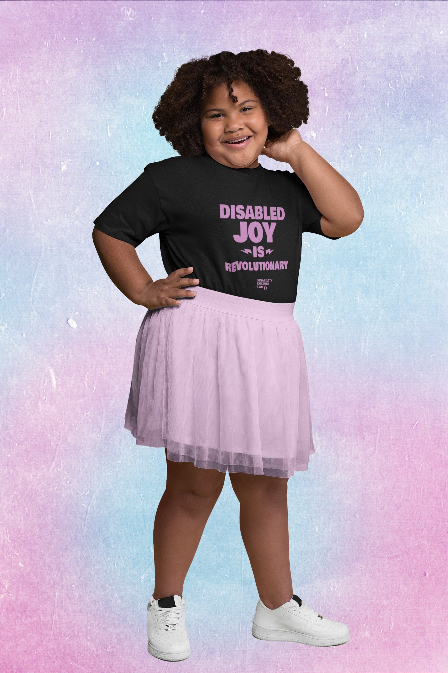 A brown-skinned young person with curly hair, wearing a purple skirt wears a black t-shirt with the words "Disabled Joy is Revolutionary."