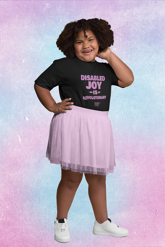 A brown-skinned young person with curly hair, wearing a purple skirt wears a black t-shirt with the words "Disabled Joy is Revolutionary."