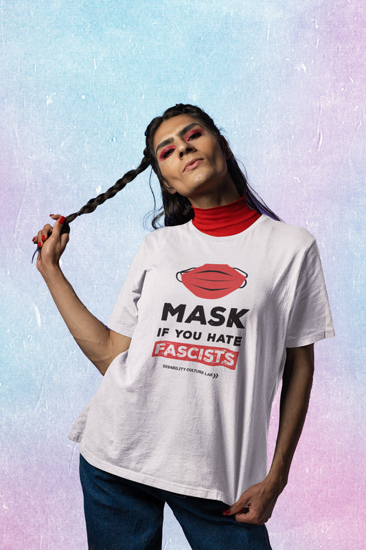 A brown-skinned trans person with long hair and fierce makeup in a white t-shirt with a red mask that reads “Mask If You Hate Fascists.” 