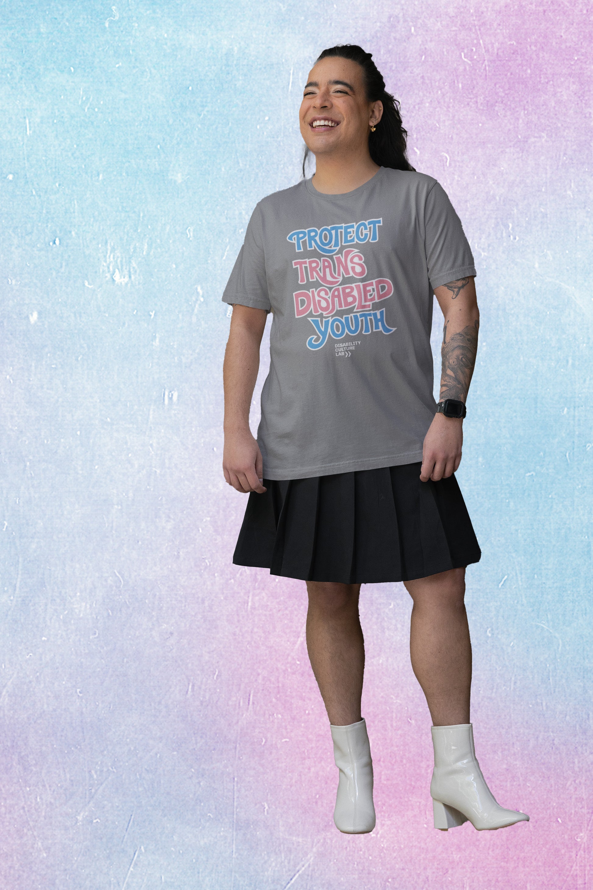 A gorgeous trans femme person in a grey t-shirt that reads “Protect Trans Disabled Youth” in hand drawn letters in the trans pride flag colors (light pink, sky blue, and white).