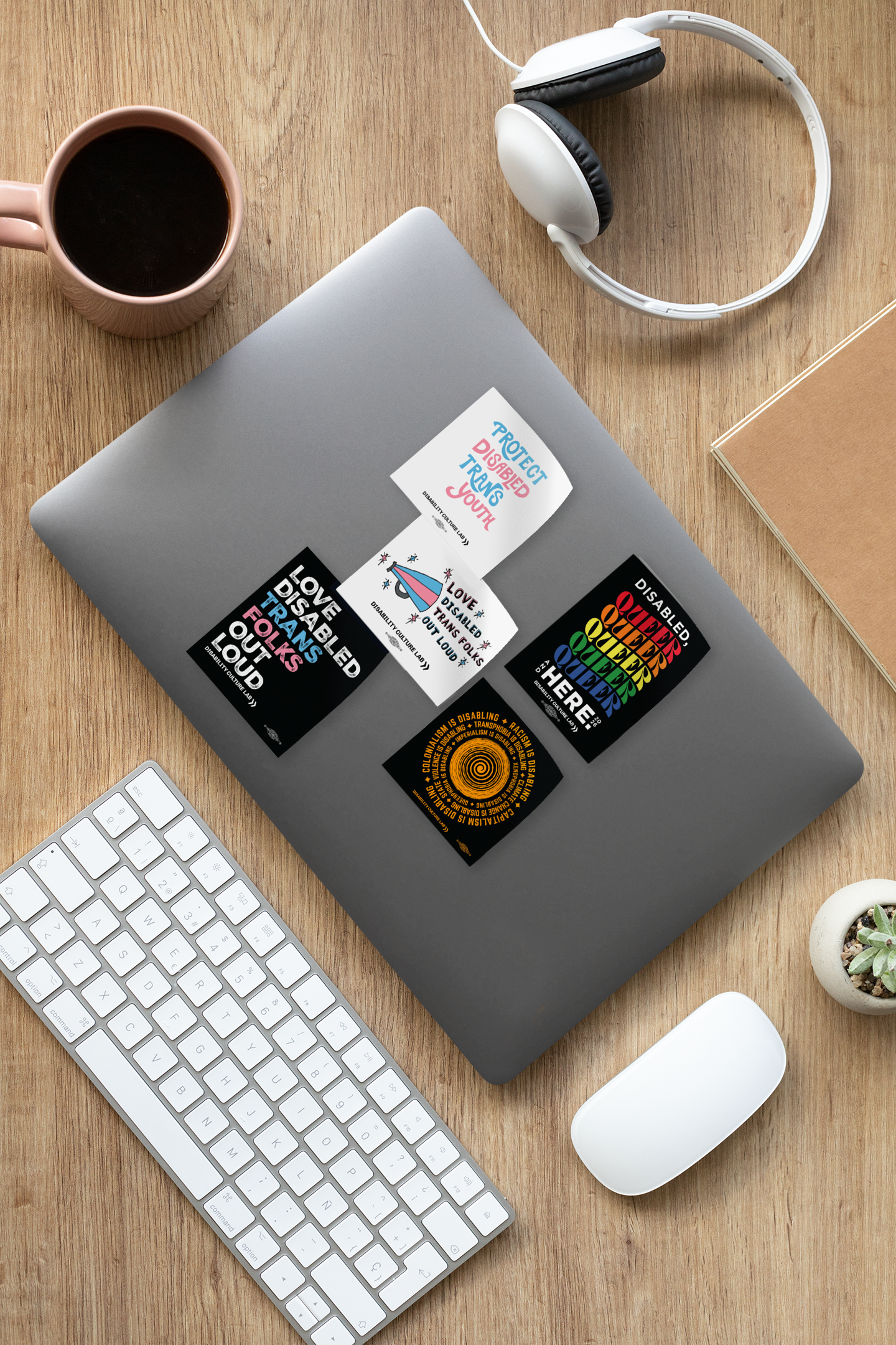 Five stickers on a gray laptop. The laptop is sitting on a wooden desk. The sticker designs are: "Protect Disabled Trans Youth." “Disabled, Queer and Here 2026” sticker is a rainbow of colors. "Love Disabled Trans Folks Out Loud" megaphone design sticker. “Oppression is Disabling” circular design sticker in yellow. "Love Disabled Trans Folks Out Loud" stacked design sticker. 