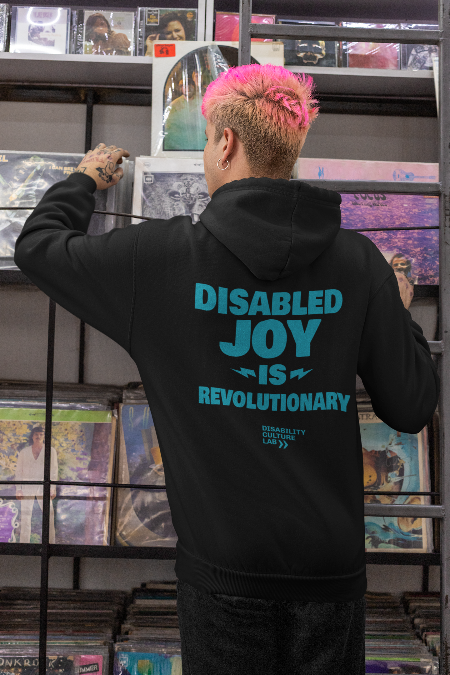 A person with short-cropped brown-blonde hair faces away from the camera while perusing records in a store, showing the back of their black zip-up hoodie that reads "Disabled Joy Is Revolutionary" in turquoise, along with the turquoise Disability Culture Lab logo.