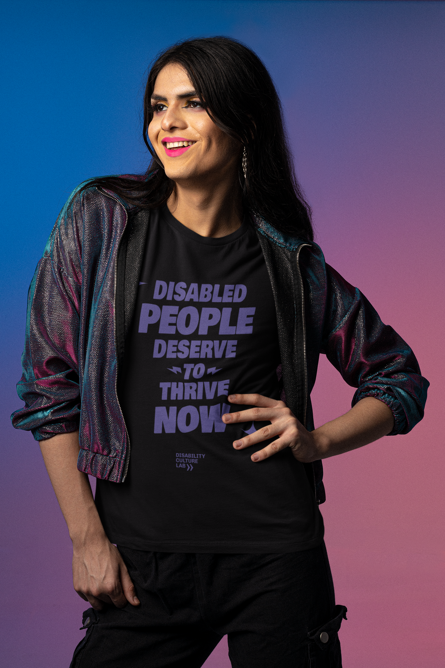 A brown-skinned trans person with long hair and fierce makeup looks right and smiles brightly. They are wearing a shimmering jacket, black pants, and a black t-shirt with the words "Disabled People Deserve To Thrive Now" in purple, with two purple sparkles, and the purple Disability Culture Lab logo below.