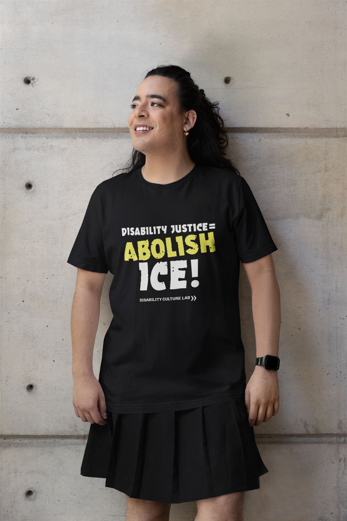A gorgeous trans femme person leans against a wall, wearing a black skirt and a black t-shirt that reads "Disability Justice = Abolish ICE!" in mostly white text, with "Abolish" in bright yellow and slightly tilted. Below is the white Disability Culture Lab logo.