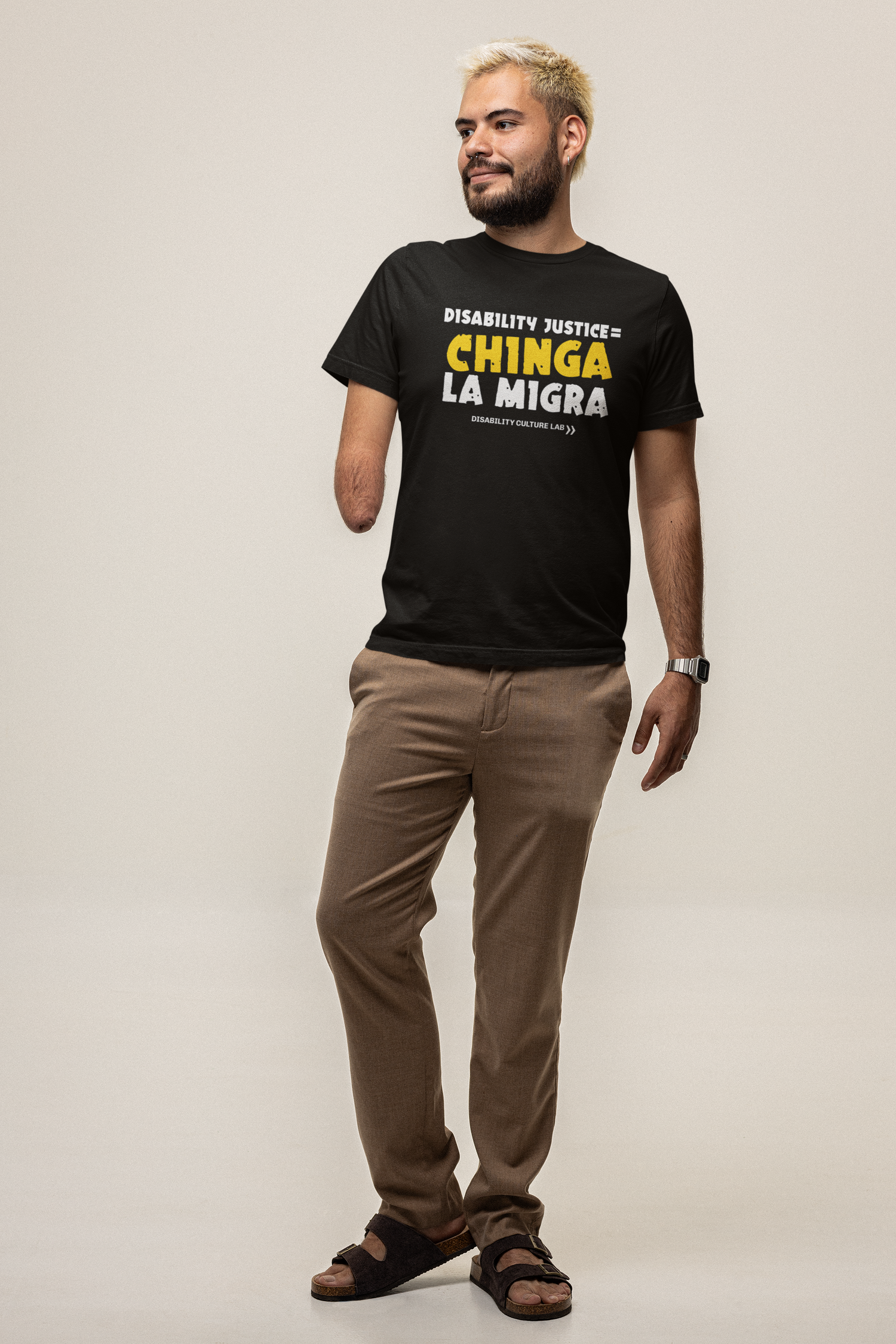 A person with blonde short-cropped hair and limb difference stands relaxed, looking to the right and smiling. They are wearing khaki pants and a black t-shirt that reads "Disability Justice = Chinga La Migra" in mostly white text, with "Chinga" in bright yellow and slightly tilted. Below is the white Disability Culture Lab logo.