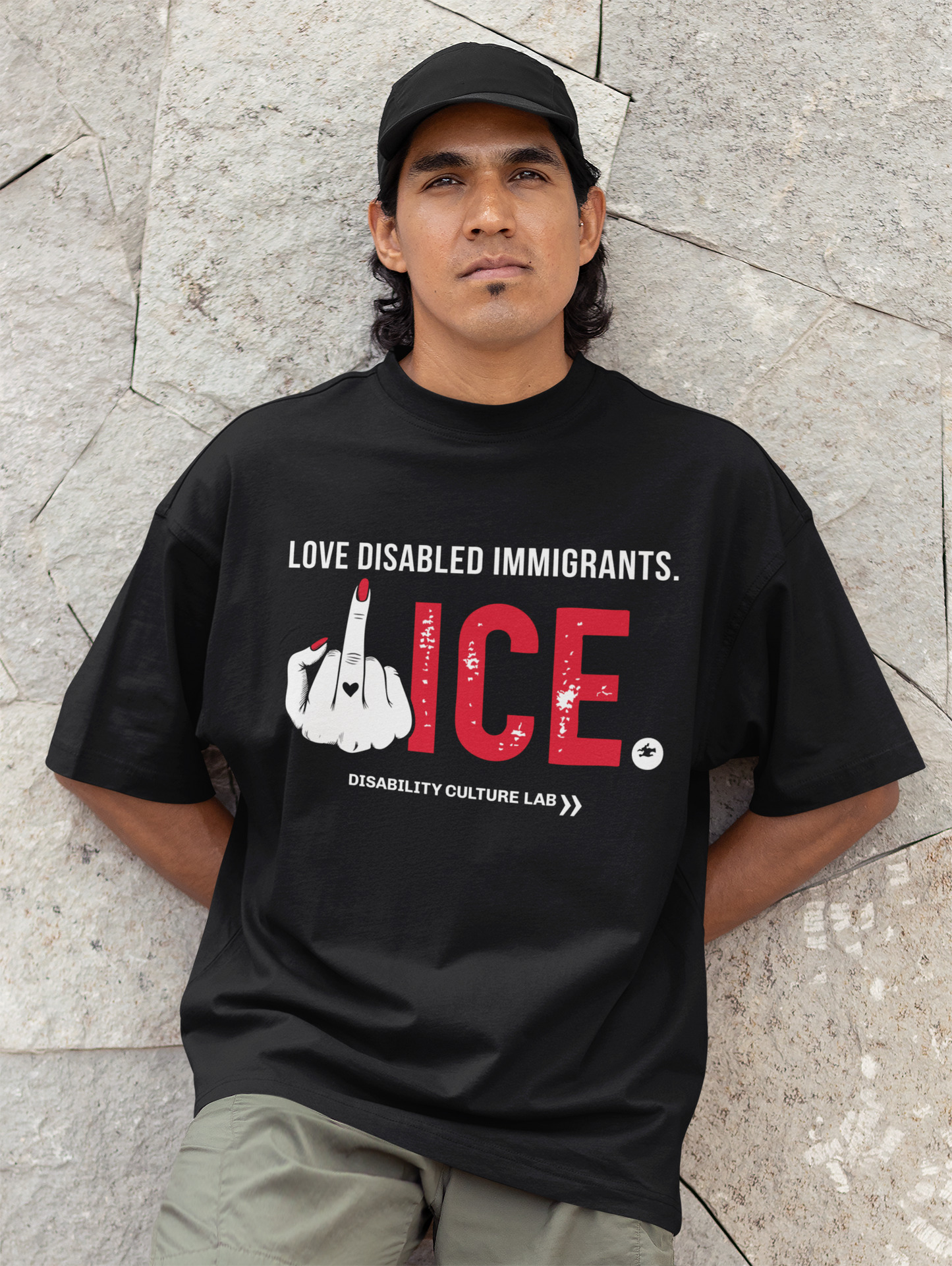 A brown-skinned person with black wavy hair leans against a wall, looking at the camera. They are wearing a black hat, gray pants, and black t-shirt that reads "Love Disabled Immigrants" in white text. Below is a hand with red nails and a black heart on the middle finger flipping the middle finger, and large textured text that reads "ICE," with "ICE" in red and the period in white. All together, the message can be read as "Love Disabled Immigrants. Fuck ICE." Below is the white Disability Culture Lab logo.