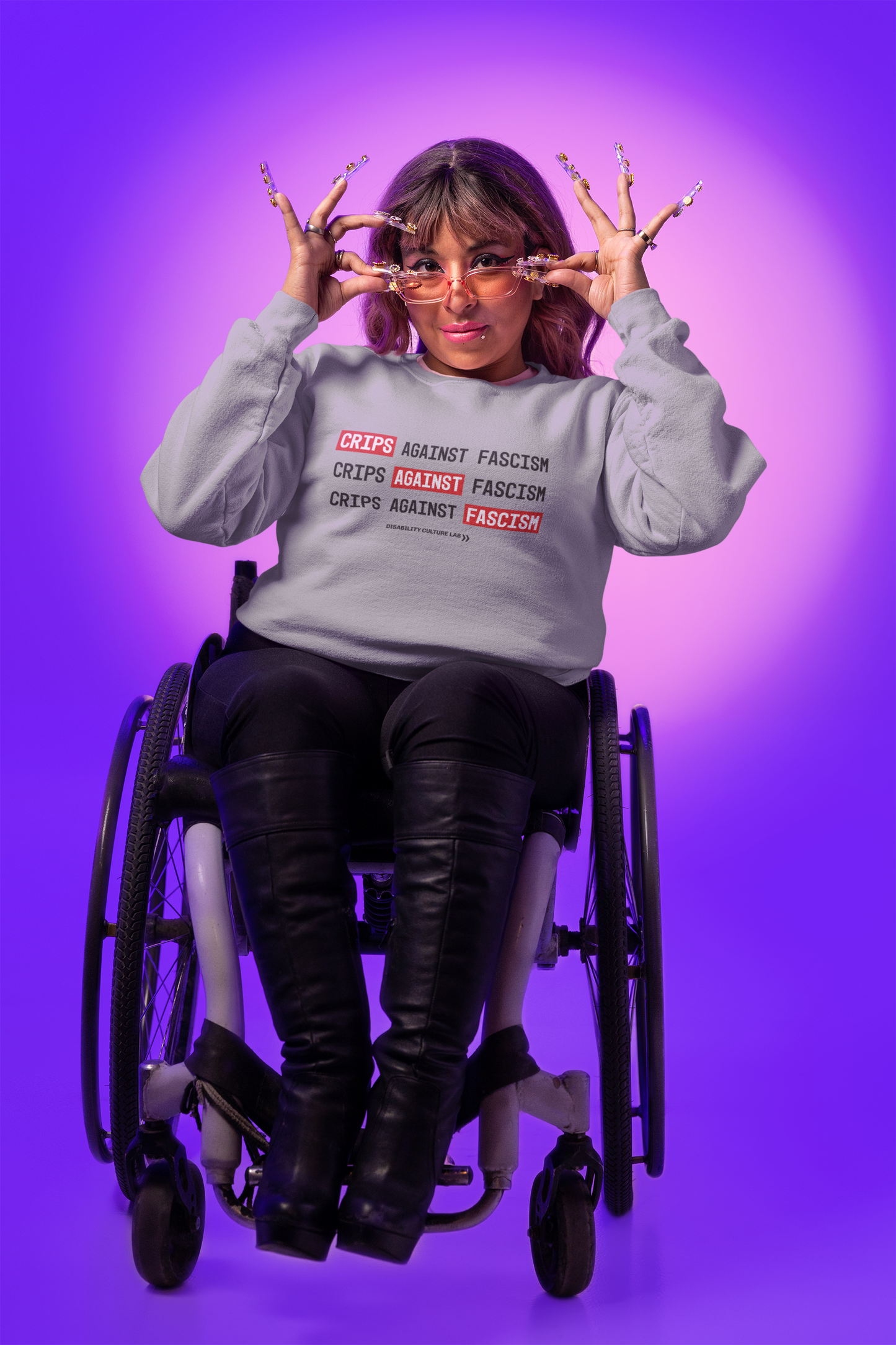 Person with brown hair, long nails, sunglasses, and winged eyeliner lowers their glasses to look at the camera. They are seated in a wheelchair, wearing black knee-high boots, black pants, and gray crewneck sweater that reads "Crips Against Fascism" three times, with "Crips" in the first line, "Against" in the second line, and "Fascism" in the third line all in white text with bright red background, so it also reads "Crips Against Fascism" diagonally. Below is the black Disability Culture Lab logo.