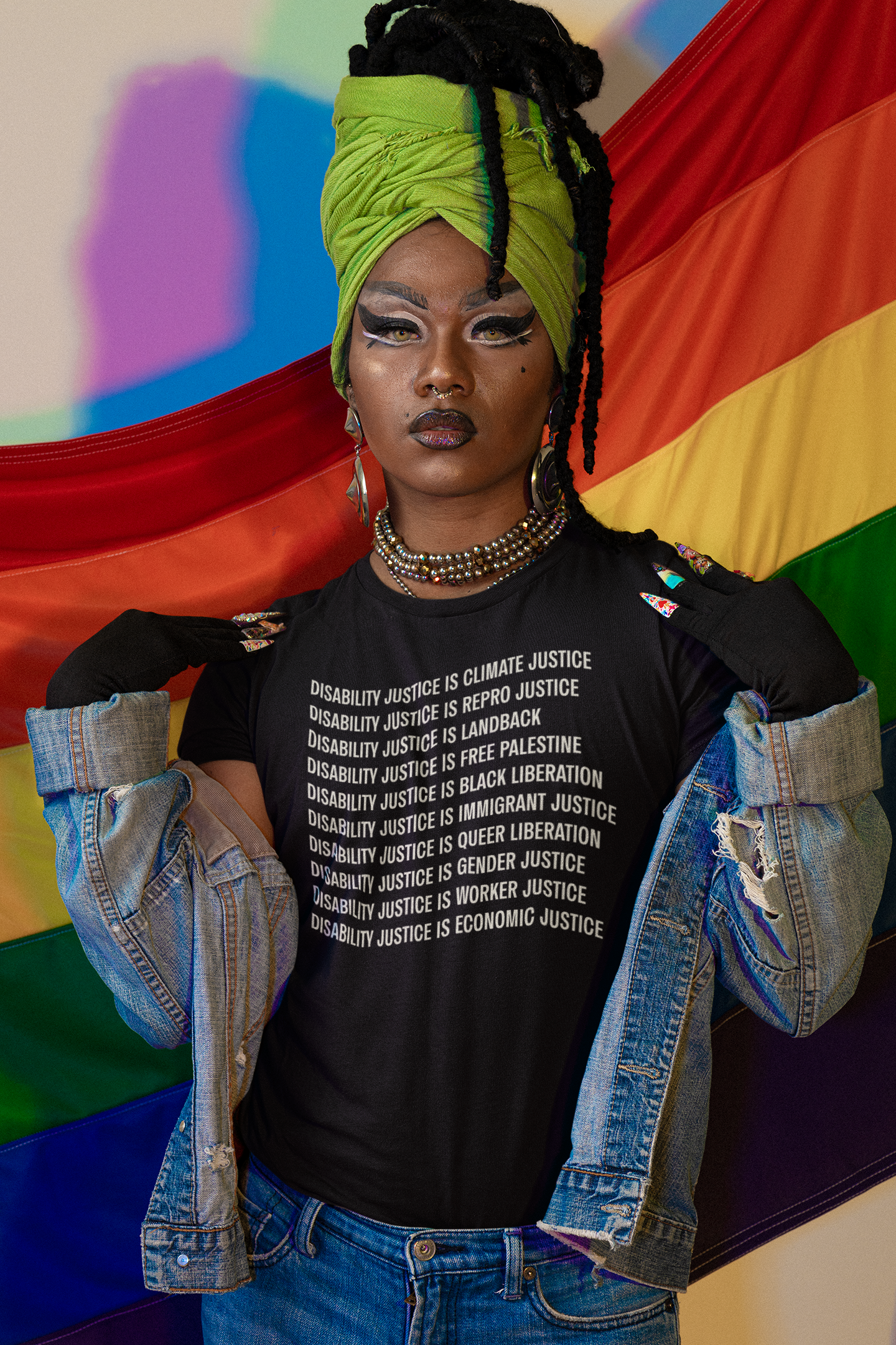 A brown-skinned person with a septum ring, dark winged eye makeup and loced hair in a green wrap, is wearing a black Disability Culture Lab shirt, Union Printed and Made in the USA, by Jennifer White-Johnson: Text reads: "Disability Justice Is... Climate Justice, Repro Justice, Landback, Free Palestine, Black Liberation, Immigrant Justice, Queer Liberation, Gender Justice, Worker Justice, Economic Justice." The background is a draped Pride flag.