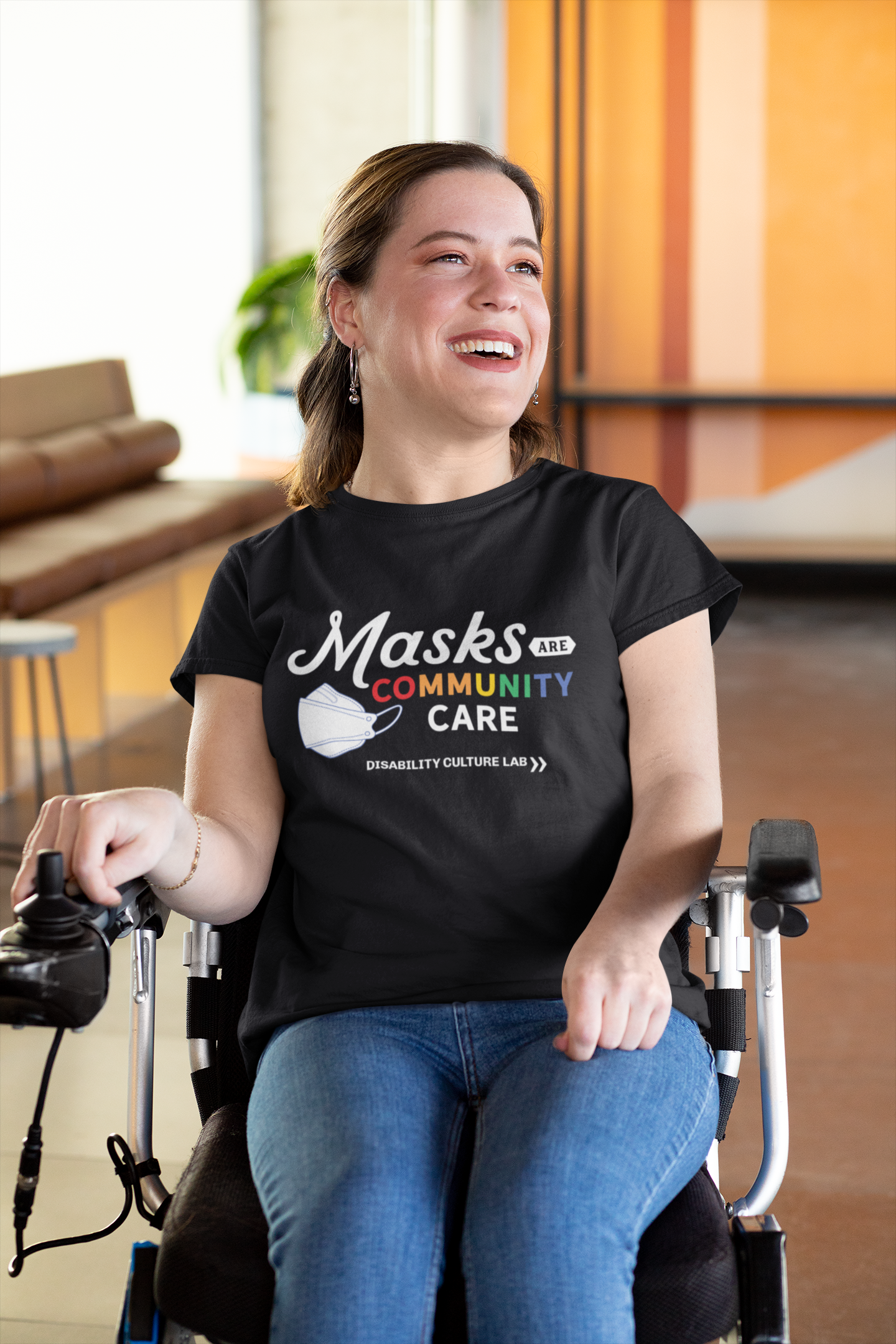 A person with brown hair, sits in a power chair, looking to the left with a bright smile. They are wearing blue jeans and a black t-shirt that reads "Masks Are Community Care," with "Masks" in a white handwritten font, "Are" in black text in a small white shape, "Community" in rainbow text, and "Care" in white text. To the left of the text is a white KF94 trifold respirator mask. Below is the white Disability Culture Lab logo.